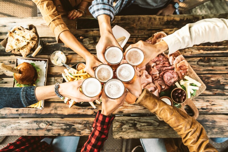 Friends,Cheering,Beer,Glasses,On,Wooden,Table,Covered,With,Delicious